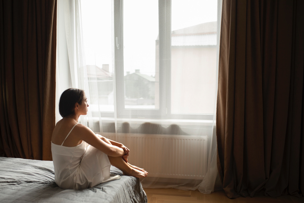 Person sitting on a bed near a window in the morning, reflecting low energy caused by irregular sleep timing